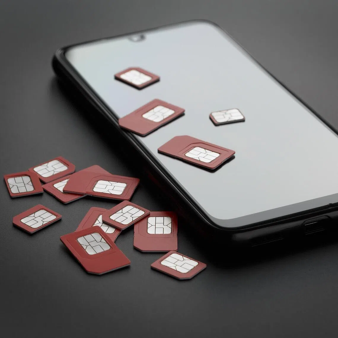 Many red SIM cards are on top of the black screen of a mobile smartphone. Mobile communication microchips and a telephone device are isolated on a white background.