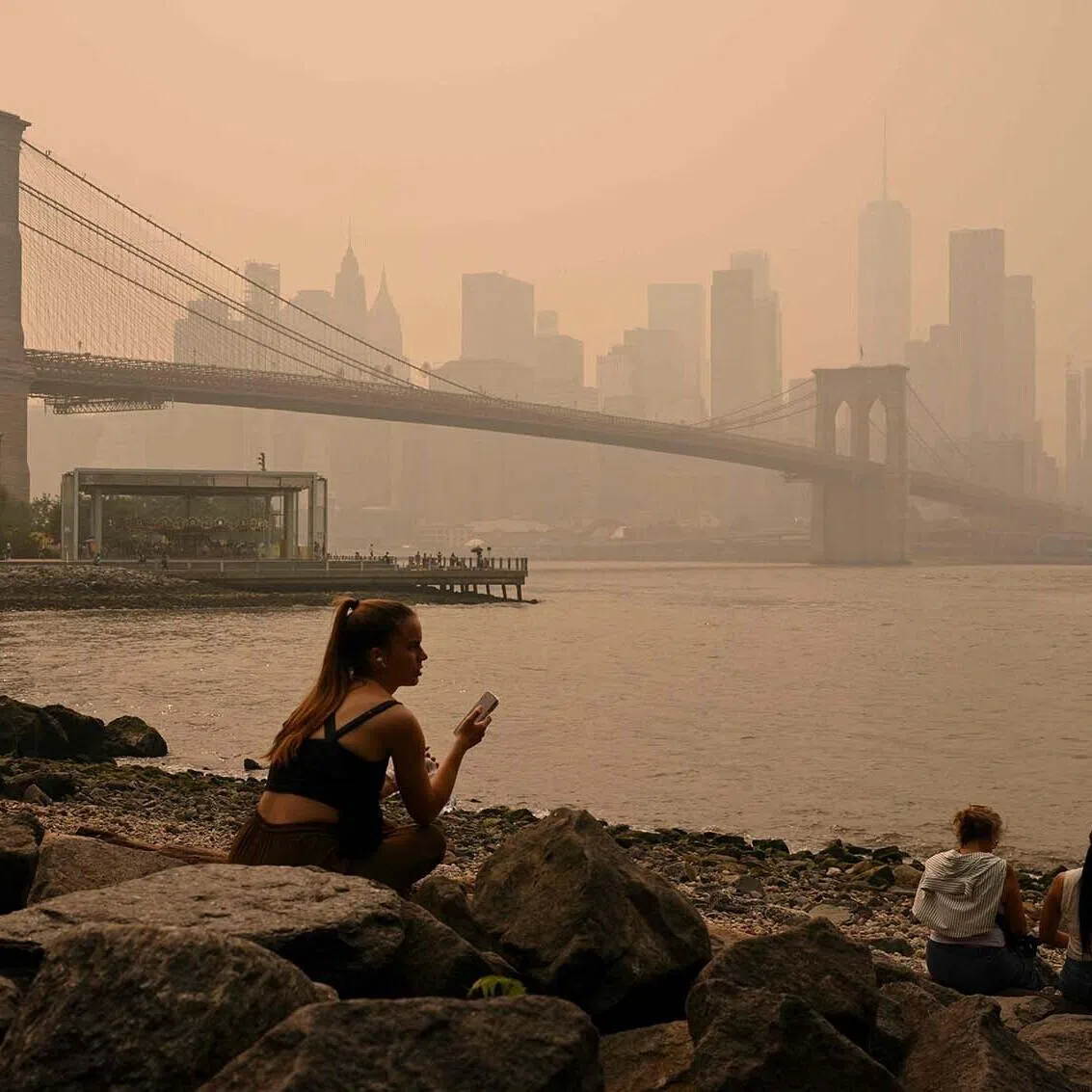 TOPSHOT - People sit near the Brooklyn Bridge as wildfire smoke from Canada causes hazy conditions on July 16, 2026 in New York. Out-of-control wildfires were raging Thursday in the Canadian province of Ontario, prompting evacuations and sending dangerous smoke billowing into the United States where millions of people were exposed to the unhealthy air. US states near the Canadian border including Minnesota, Wisconsin, Michigan and Illinois were particularly choked, as forecasters predicted the Northeast including New York would also see air quality continue to deteriorate. (Photo by ANGELA WEISS / AFP)