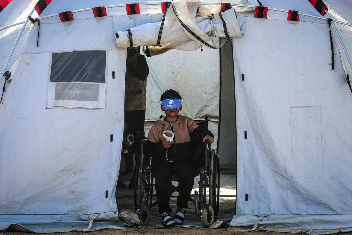 TOPSHOT - A Palestinian child sitting in a wheelchair wearing goggles and holding a joy stick experiences virtual reality as a medical technology support team launches an initiative in the city of Al-Zawayda, in the central Gaza Strip on November 30, 2025, featuring therapy sessions using virtual reality technology, introduced due to the two-year-old war that was sparked by the Palestinian militant group Hamas's October 7, 2023 attack on Israel, and Israel's retaliatory assault on the Gaza Strip. Children, traumatised by war have been given virtual reality headsets, to transport them from scenes of destruction to green gardens, tranquil beaches, and cities brimming with games, with the aim of helping to alleviate the psychological trauma they endure daily. One of the medical technology team members said the idea stemmed from the need to "change the harsh reality that children live in, even if only for a few hours, by giving them a chance to feel safe and joyful." These sessions rely on programs specifically designed for traumatised children, taking into account their physical and psychological condition and helping them rebuild positive perceptions of the world. (Photo by Eyad Baba / AFP)