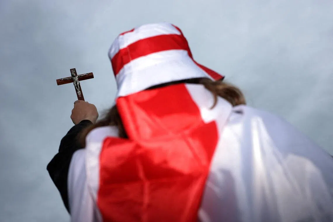 A person in a red and white robe holding a cross against a cloudy sky.