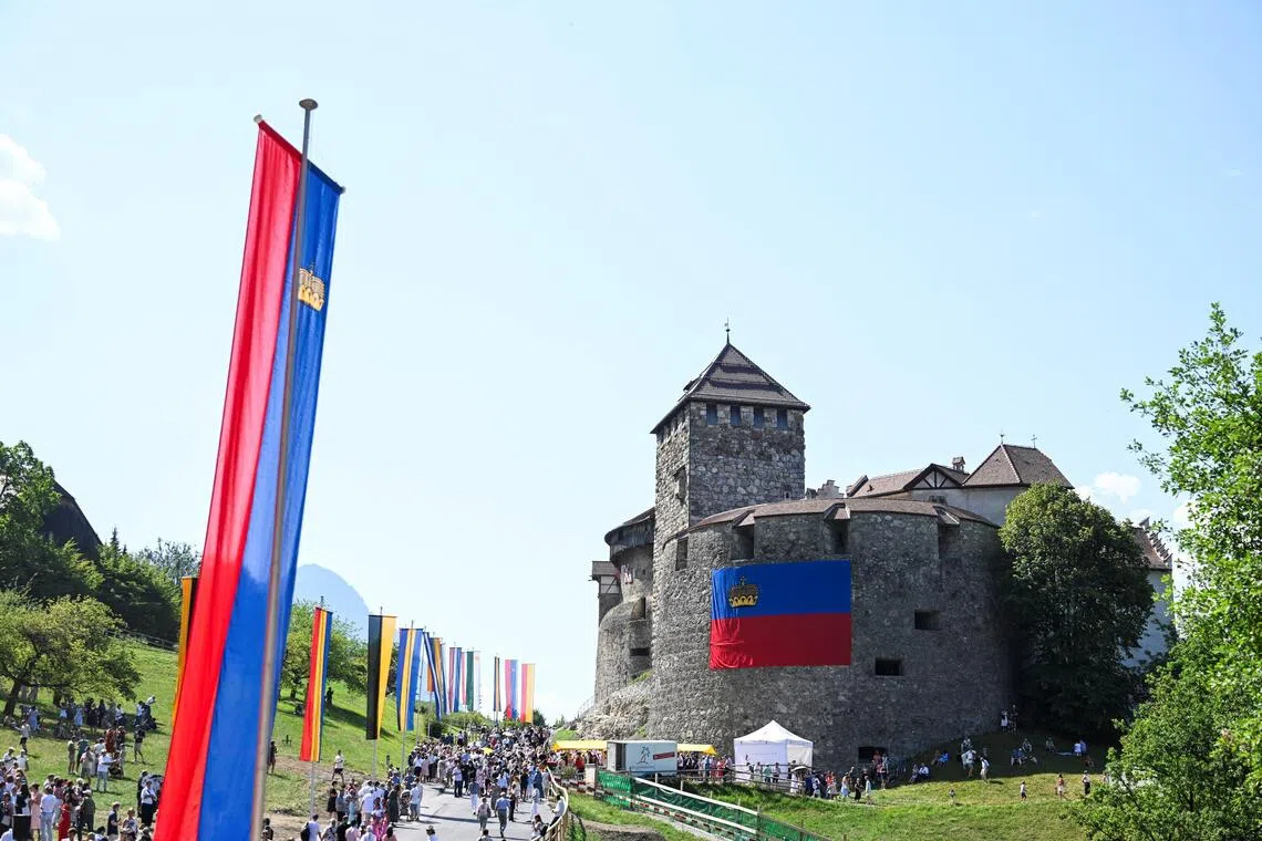 A crowd of people walking past flags towards a stone castle on a hillside.