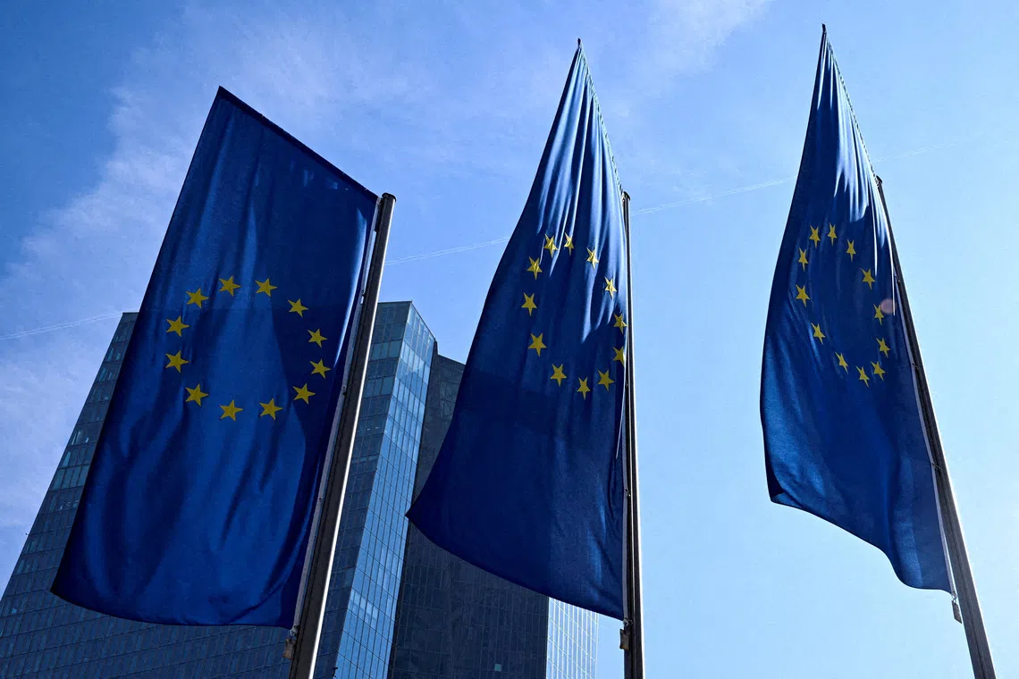 Three European Union flags flying in front of a glass building.