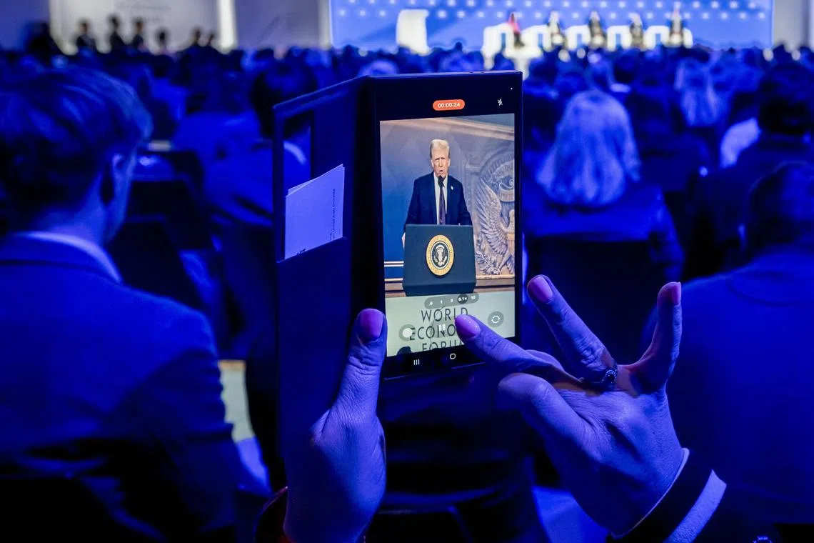 A woman films with her mobile as US President Donald Trump is seen on a giant screen during his address by video conference at the World Economic Forum (WEF) annual meeting in Davos on January 23, 2025. (Photo by Fabrice COFFRINI / AFP)