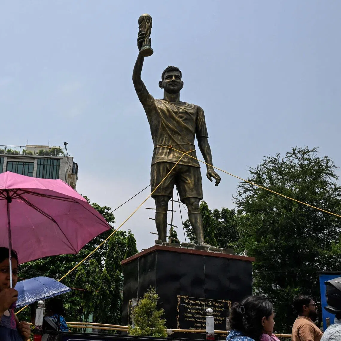 Commuters walk past a statue of Argentine football player Lionel Messi that has been tied with rope for stability from rain and thunderstorms, prior to its re-installation in a safer place, in Kolkata on May 27, 2026. Messi inaugurated the statue virtually during his visit to the city in December 2025.
