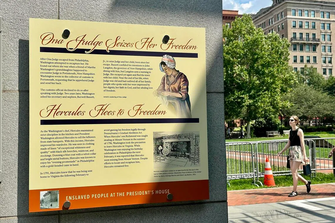 A historical sign about enslaved people at the President's House stands next to a sidewalk where a woman is walking.