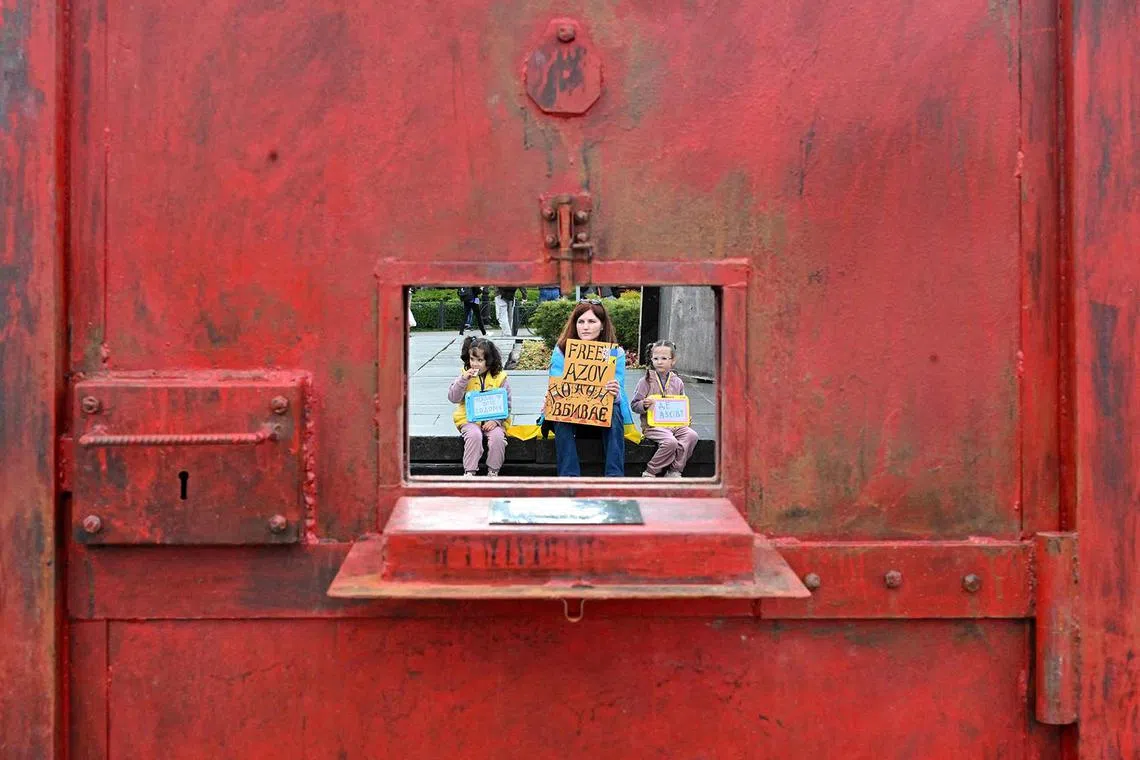 A protester and two children holding placards as they are seen through a window of a "door to a prison cell" installation during a rally marking the third anniversary of the capture of Azov Brigade fighters in Mariupol and to call to speed up exchange of the Ukrainian prisoners of war of the brigade and from the other units, in Kyiv on May 18, 2025, amid Russian invasion in Ukraine. 