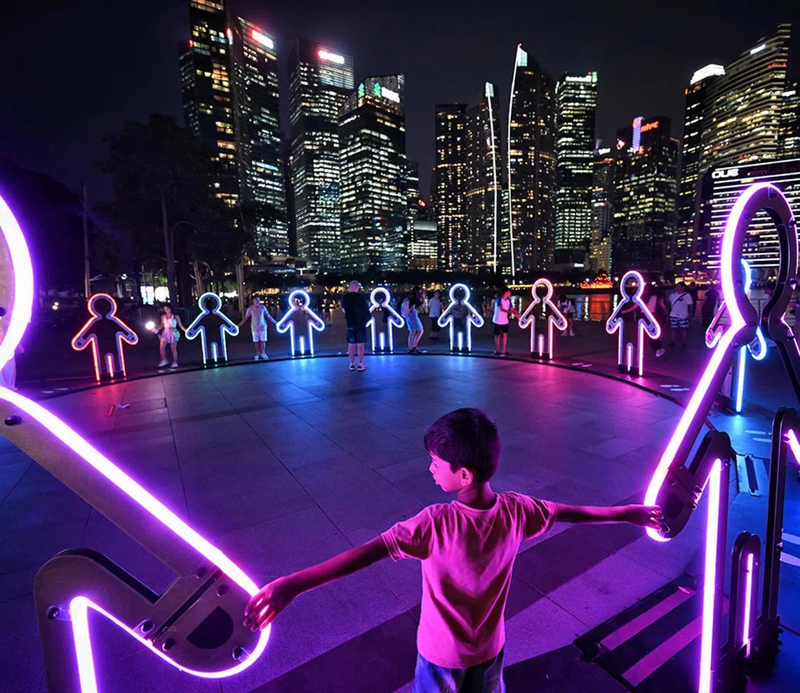 (SUGGESTED MAIN PIC #2) Embrace by Beamhacker (Australia), presented by Defence Collective Singapore at Event Square, features life-sized silhouettes inspired by children's paper chains. When visitors hold the hands of the figures, ripples of light and sound are triggered, culminating in a full symphony when the circle is completed. The installation evokes the joy of childhood play while symbolising unity between Singapore’s military and civilians. The silhouettes, adorned with camouflage patterns representing the four SAF services, stand as a tribute to collective strength. A commissioned score by Peter McAvan accompanies the experience.