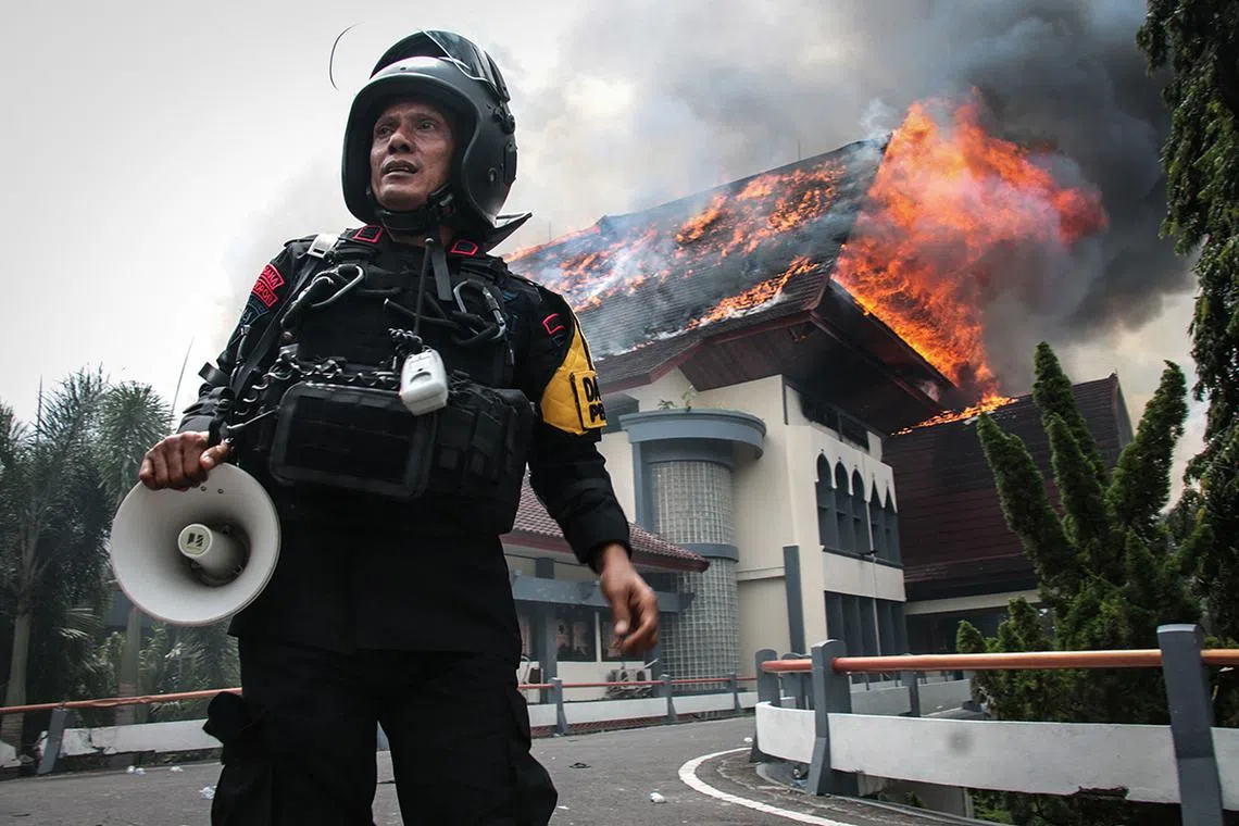 TOPSHOT - A police officer stands with a megaphone as the building of the West Nusa Tenggara Provincial Council office burns after being set on fire by protesters in Mataram on Lombok Island, West Nusa Tenggara, on August 30, 2025. Indonesia was rocked by protests in major cities including the capital Jakarta on August 29 after footage spread of a motorcycle taxi driver being run over by a police tactical vehicle in earlier rallies against low wages and financial perks for lawmakers. (Photo by Sadim / AFP)