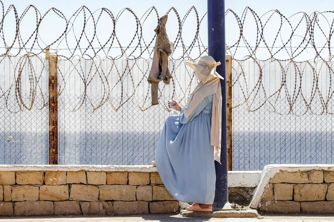 TOPSHOT - A woman sits by a barbed-wire fence as relatives and friends wait for the return of people who had previously crossed into Spain's North African enclave of Ceuta, at Morocca's Bab Sebta border crossing on the outskirts of Fnideq on August 2, 2026. As many as 60,000 migrants entered Ceuta in recent days, mostly swimming around the small border post that juts into the Mediterranean Sea, in an unprecedented surge that prompted an international crisis for Spain. The mass crossing into the tiny Spanish territory resulted in at least 67 deaths, according to Spanish authorities, and led to clashes with Moroccan police on the shared border. (Photo by Abdel Majid BZIOUAT / AFP)