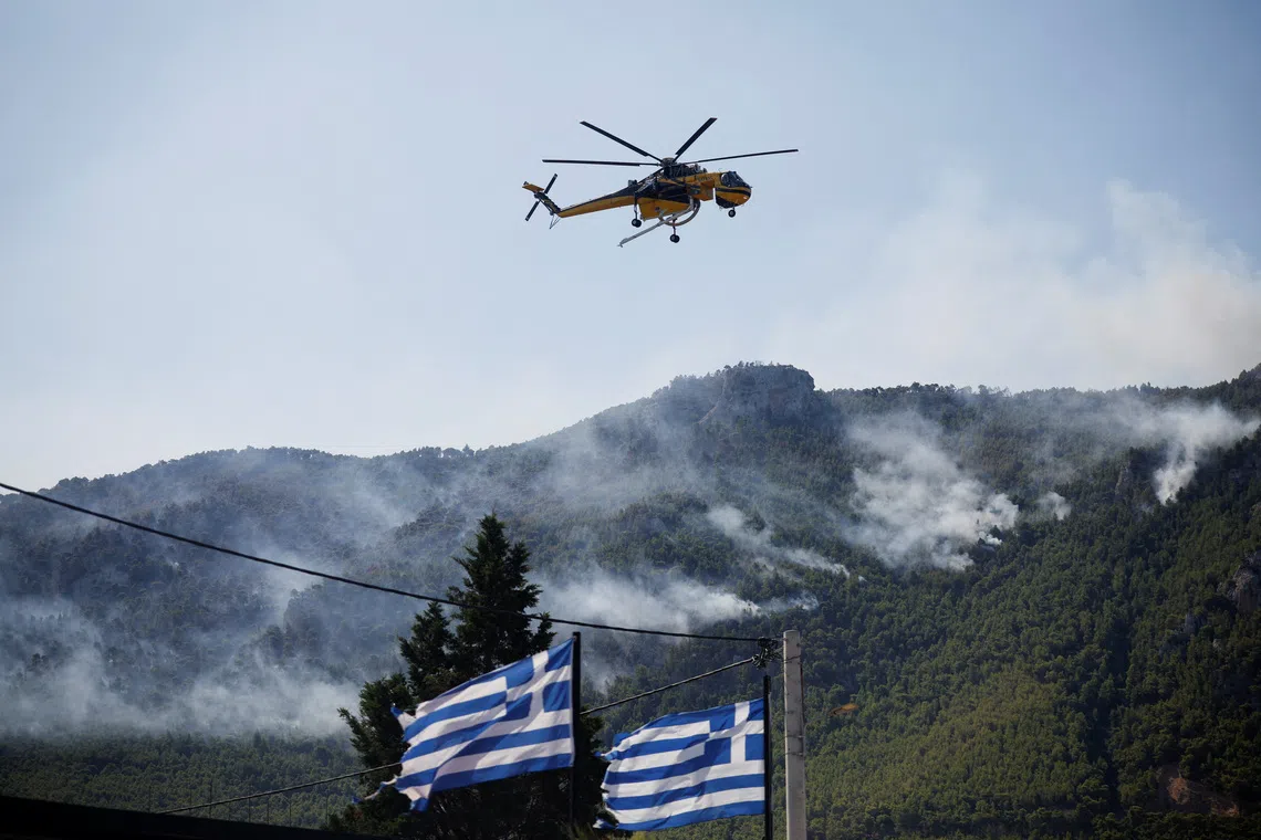 A yellow helicopter flies over a smoking mountain forest with two Greek flags in the foreground.