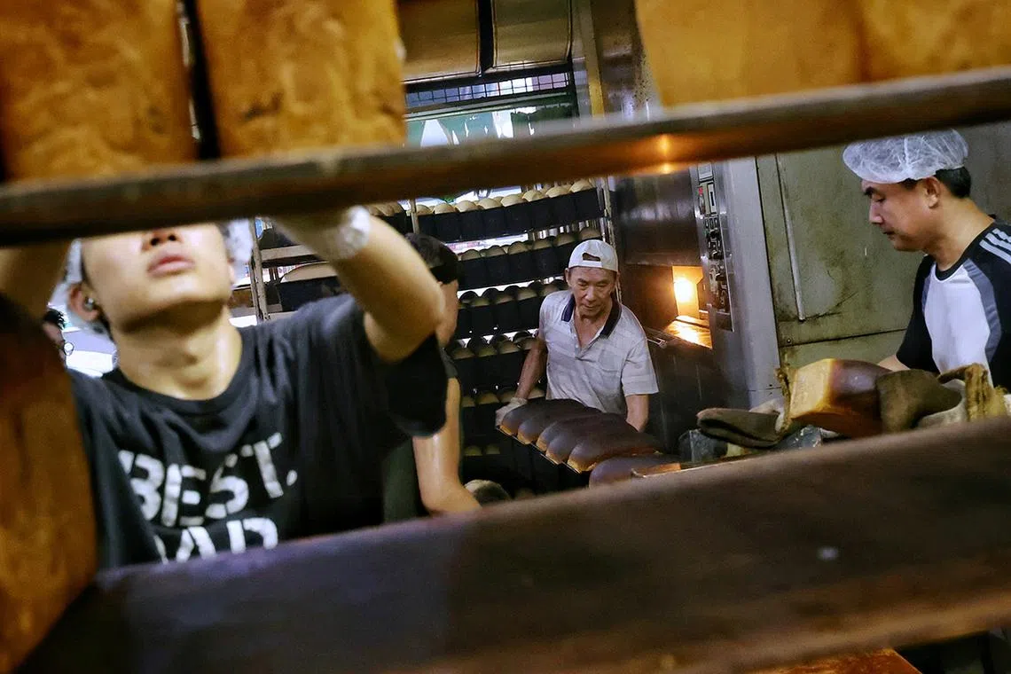 12. (SUGGESTED MAIN PIC 1) Senior baker Goh Wah Heng (centre), 72, taking out baking tins of traditional bread loaves from the revolving oven for his fellow bakers to load onto a rack to cool in the kitchen of Sweetlands Confectionery and Bakery in Kim Keat Lane on May 15, 2025. The bakery can produce more than 1,000 loaves of traditional bread in a day.