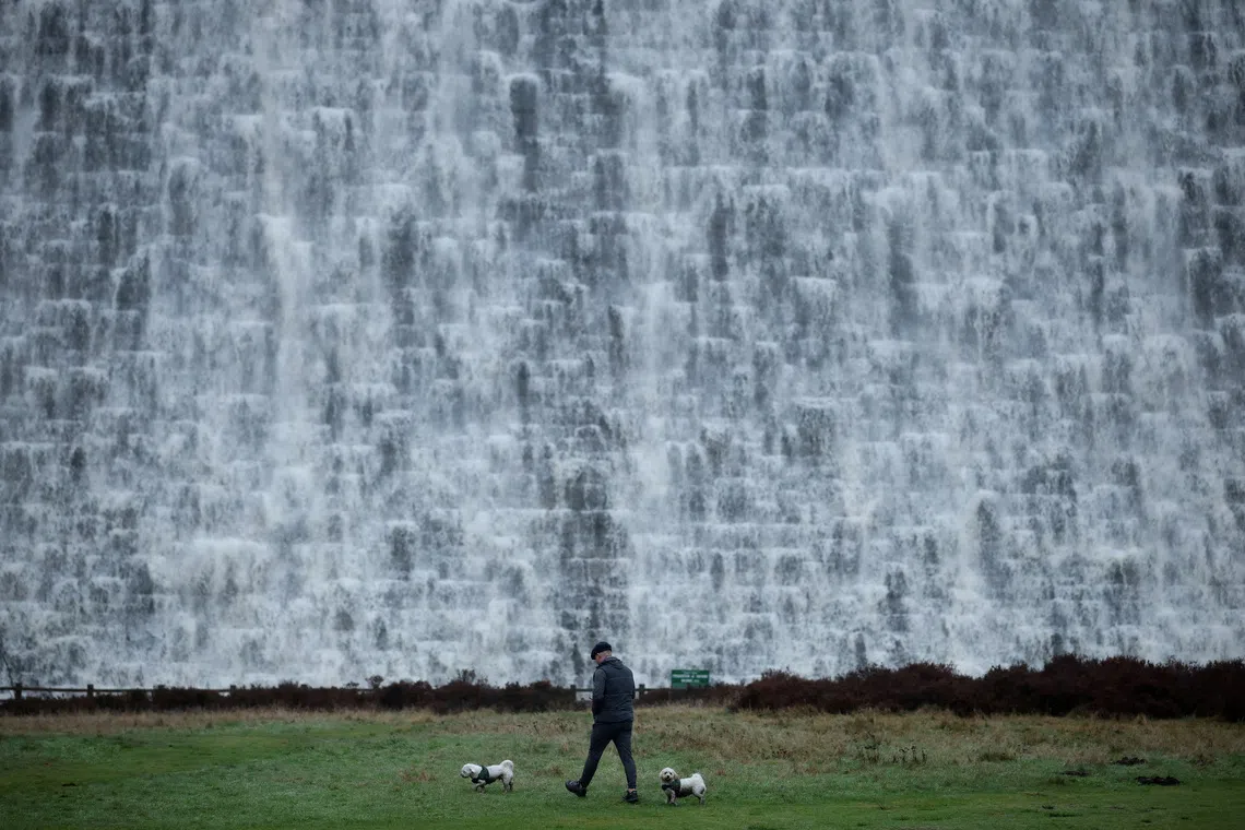 A man walking his dogs in front of the wall of the Derwent Dam as water spills over the top following a prolonged period of rain, near Edale, Britain, on Dec 7, 2025. 