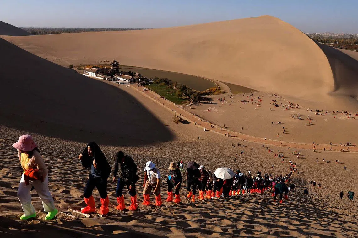 Tourists wearing shoe protectors climb up a dune with the overlook view of Crescent Spring at Mingsha Mountain, during an organised media tour to Dunhuang, in Gansu province, China October 15, 2024. REUTERS/Tingshu Wang TPX IMAGES OF THE DAY