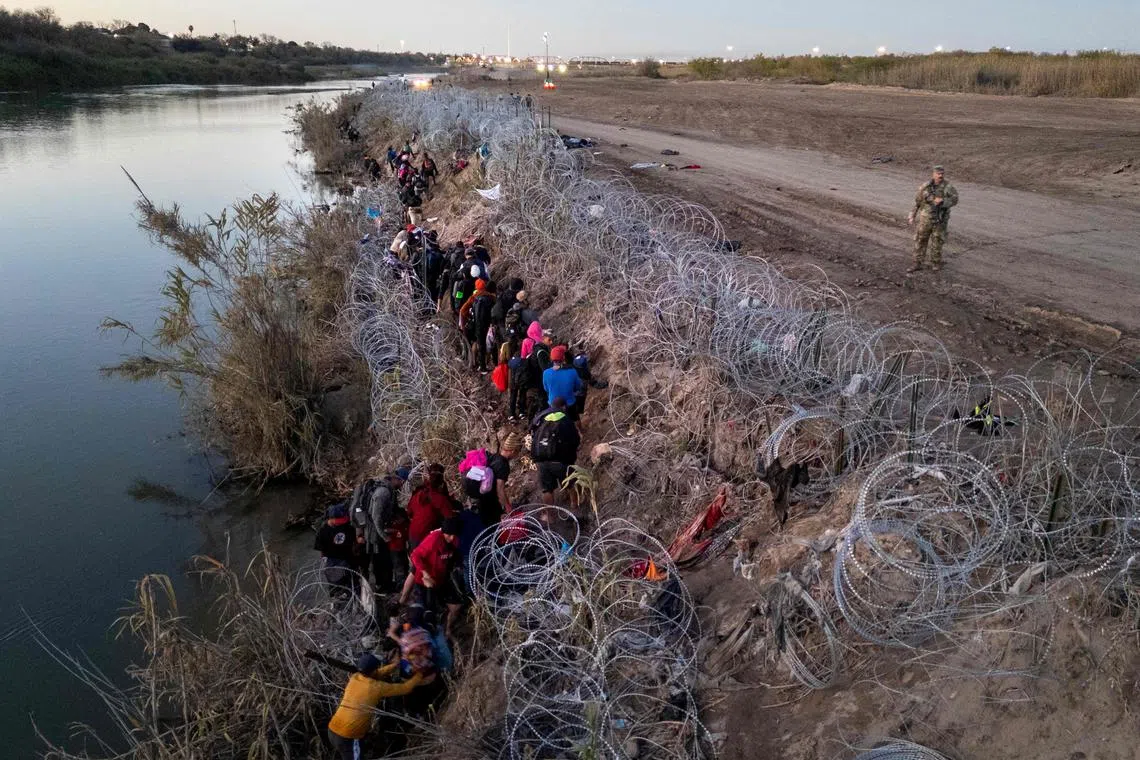 Migrants picking their way through razor wire after crossing the Rio Grande into the United States on Dec 17, 2023, in Eagle Pass, Texas. A surge of migrants, as many as 12,000 per day, crossing the U.S. southern border has overwhelmed U.S. immigration authorities in recent weeks