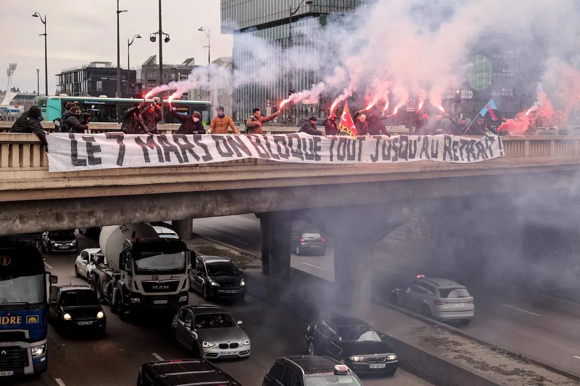 TOPSHOT - Bus drivers of State-owned RATP, which operates the greater Paris transport system, burn flares behind a banner reading in French "on March 7, let's block everything until withdrawal" on a bridge above Paris' ring road "Le Peripherique" in Paris on March 6, 2023, as massive strikes are expected from March 7 in France against French government's proposed pension reform, with unions promising to bring the country "to a standstill" and strikes set to hit many sectors such as transport, energy and oil refining. (Photo by Thomas SAMSON / AFP)