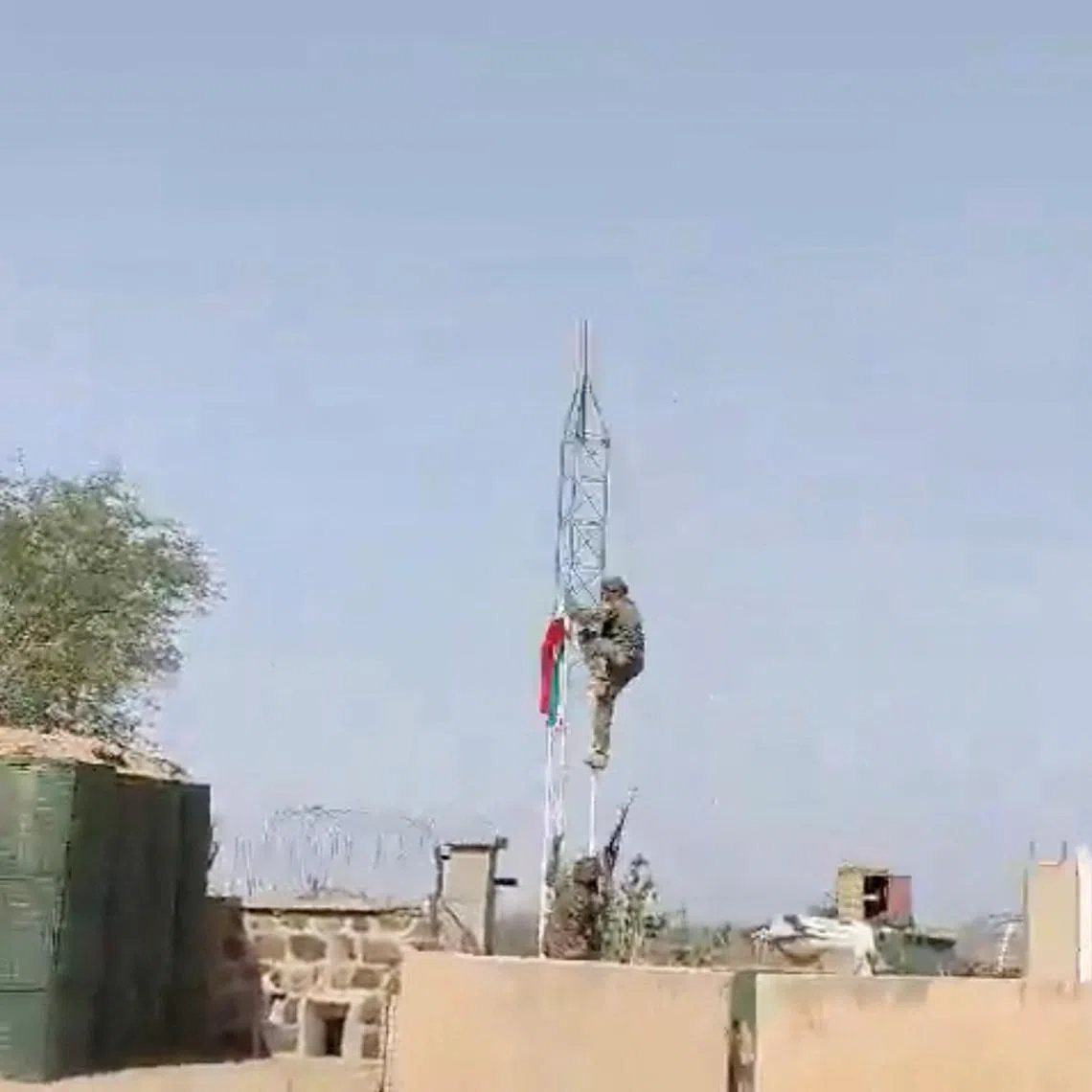 A militant climbs up a tower with a flag at at what appears to be a military base with barricades and makeshift container buildings, in Tessalit, Mali, in this screen grab obtained from a social media video released on May 1, 2026. SOCIAL MEDIA/via REUTERS
