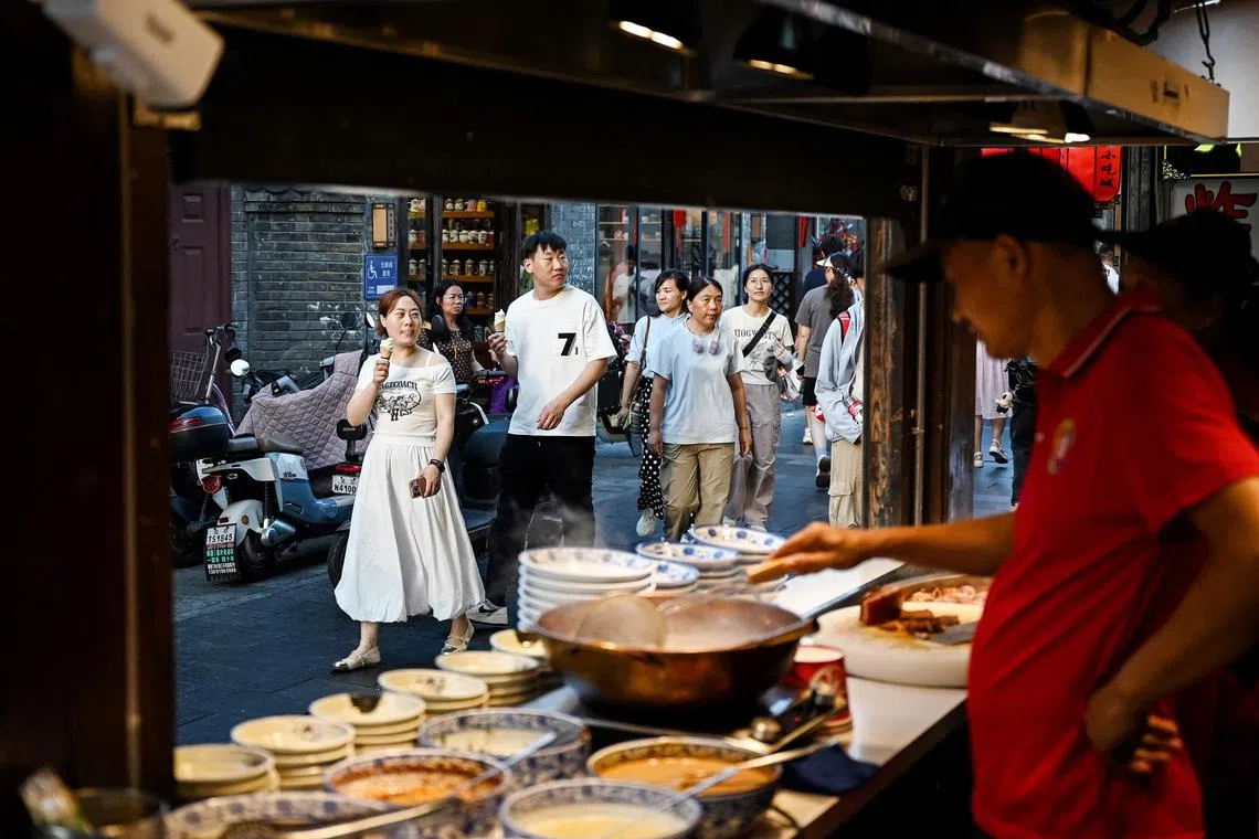 People walk past a food vendor cooking traditional snacks along an alley near the Houhai Lake in Beijing on July 22, 2025. (Photo by Greg Baker / AFP)