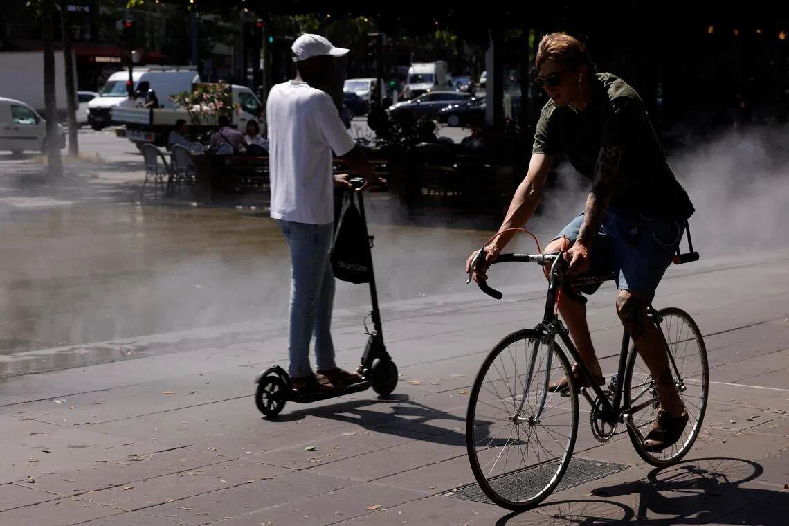 Two men, one on a scooter and one on a bicycle, are on a wet city sidewalk and street.