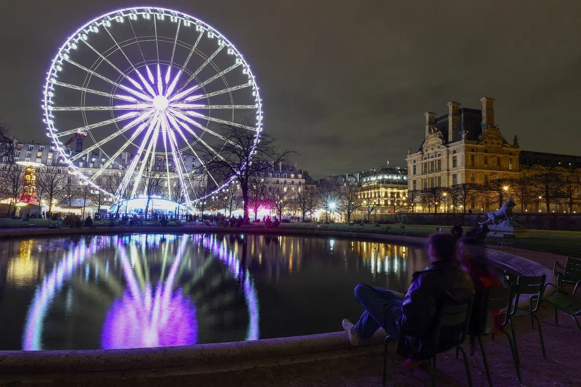 A couple sitting by a pond as the Grande Roue de Paris lights up, with the Louvre Museum seen in the background, as Paris marks the start of its Christmas holiday lights season, in France, on Dec 3, 2025. 