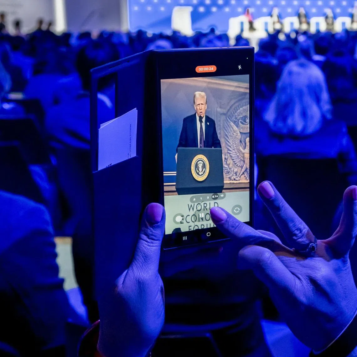 A woman films with her mobile as US President Donald Trump is seen on a giant screen during his address by video conference at the World Economic Forum (WEF) annual meeting in Davos on January 23, 2025. (Photo by Fabrice COFFRINI / AFP)