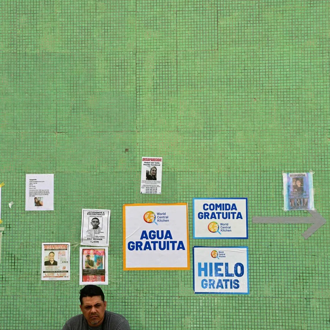 A man sits against a wall displaying signs with pictures and data of missing people and ads offering free food, ice, and water, in Caraballeda, state of La Guaira, Venezuela, on July 10.