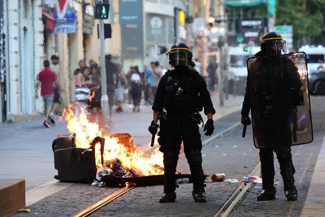 TOPSHOT - French riot police officers stand guard next to a burnt out trash bin during a demonstration against police in Marseille, southern France on July 1, 2023, after a fourth consecutive night of rioting in France over the killing of a teenager by police. French police arrested 1311 people nationwide during a fourth consecutive night of rioting over the killing of a teenager by police, the interior ministry said on July 1, 2023. France had deployed 45,000 officers overnight backed by light armoured vehicles and crack police units to quell the violence over the death of 17-year-old Nahel, killed during a traffic stop in a Paris suburb on June 27, 2023. (Photo by CLEMENT MAHOUDEAU / AFP)