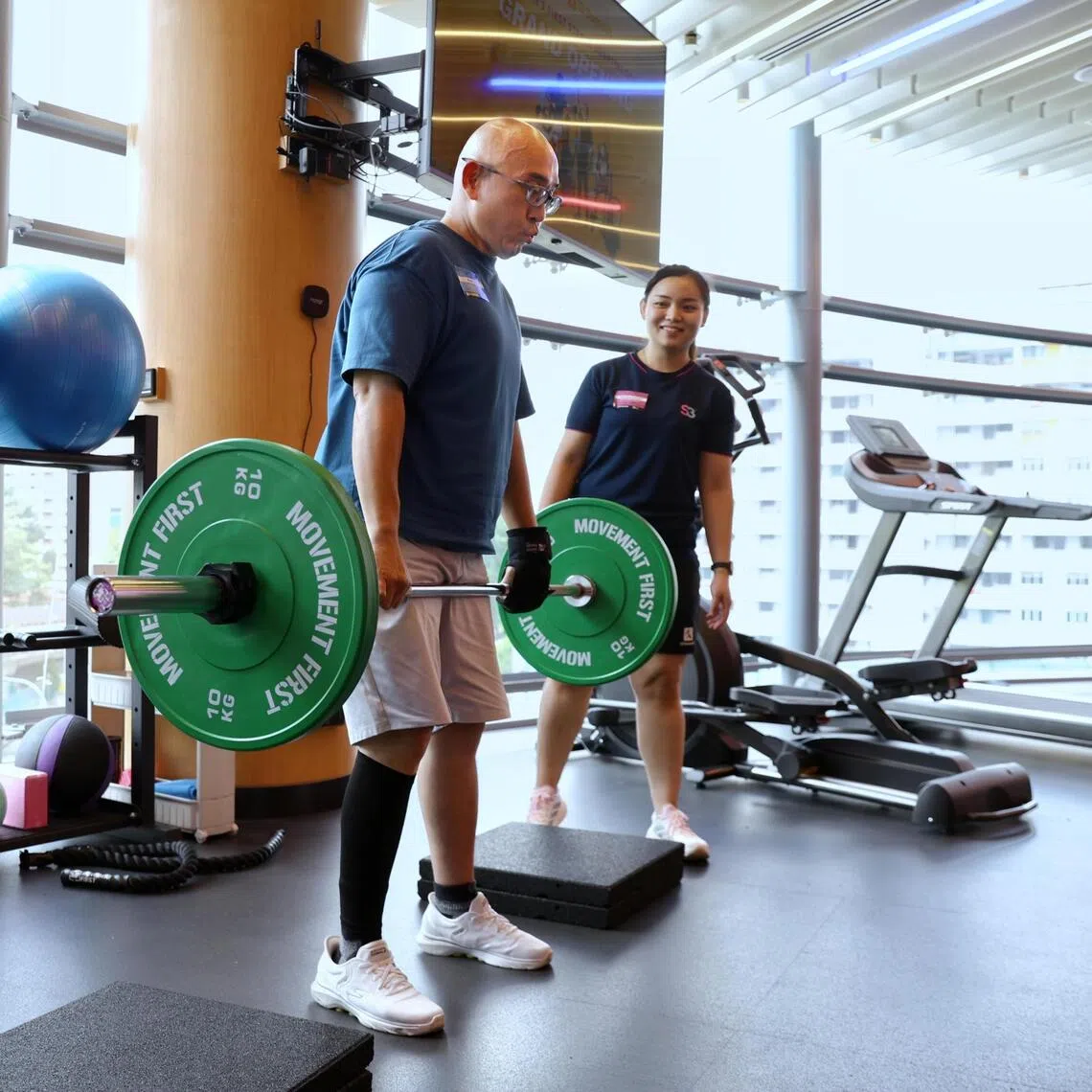 Stroke survivor Edmond Poh (left) using an assistive hand grip on his left hand while lifting weights under the watchful eyes of Exercise trainer Reosherra Lim. A year ago, he was could not have done this as he still needed a cane while walking.