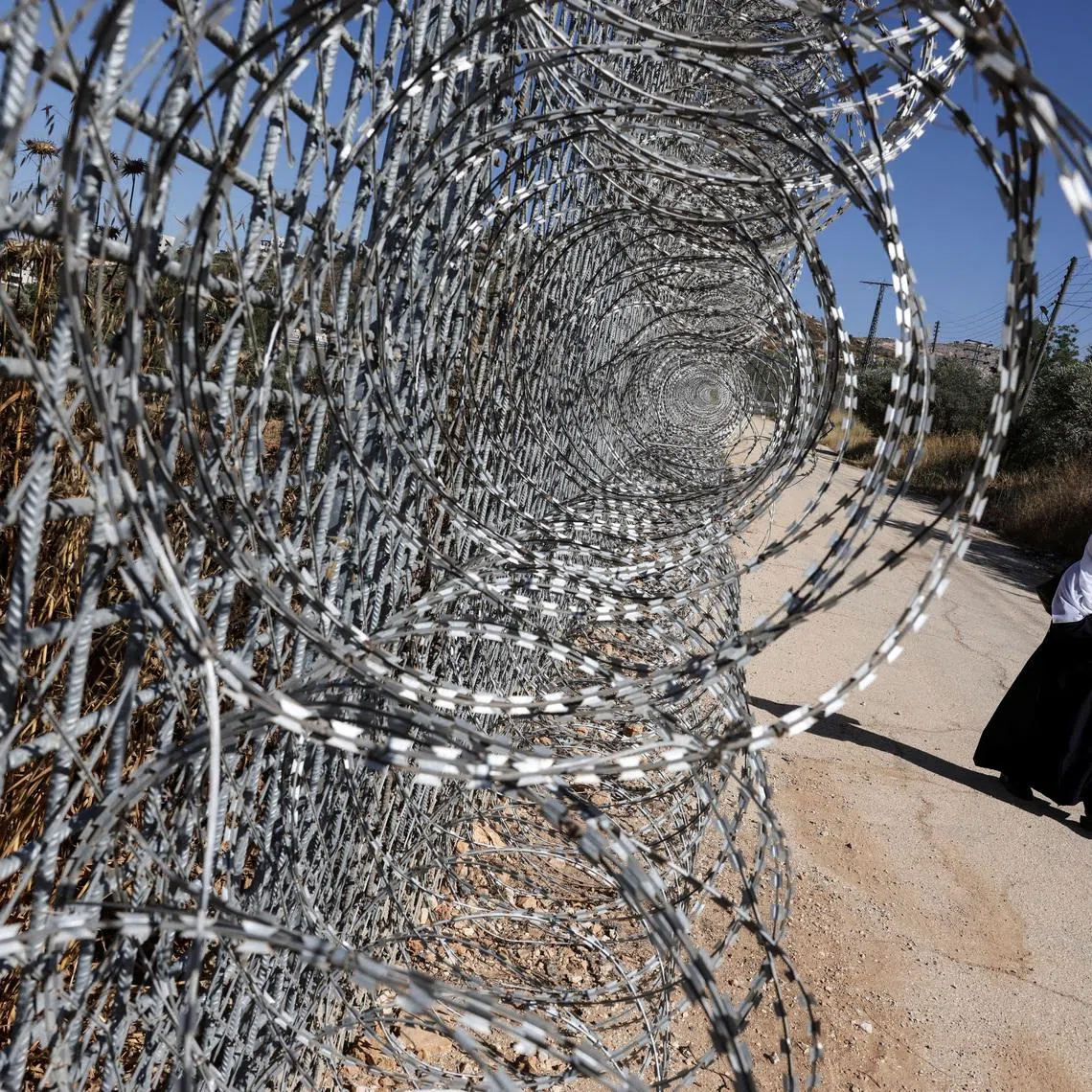 Jawaher Foqahaa walks past the tall metal fencing installed by the Israeli military that closed off four of the five entrances to their town and runs directly past the family home, in Sinjil, near Ramallah, in the Israeli-occupied West Bank, June 26, 2026. REUTERS/Ammar Awad