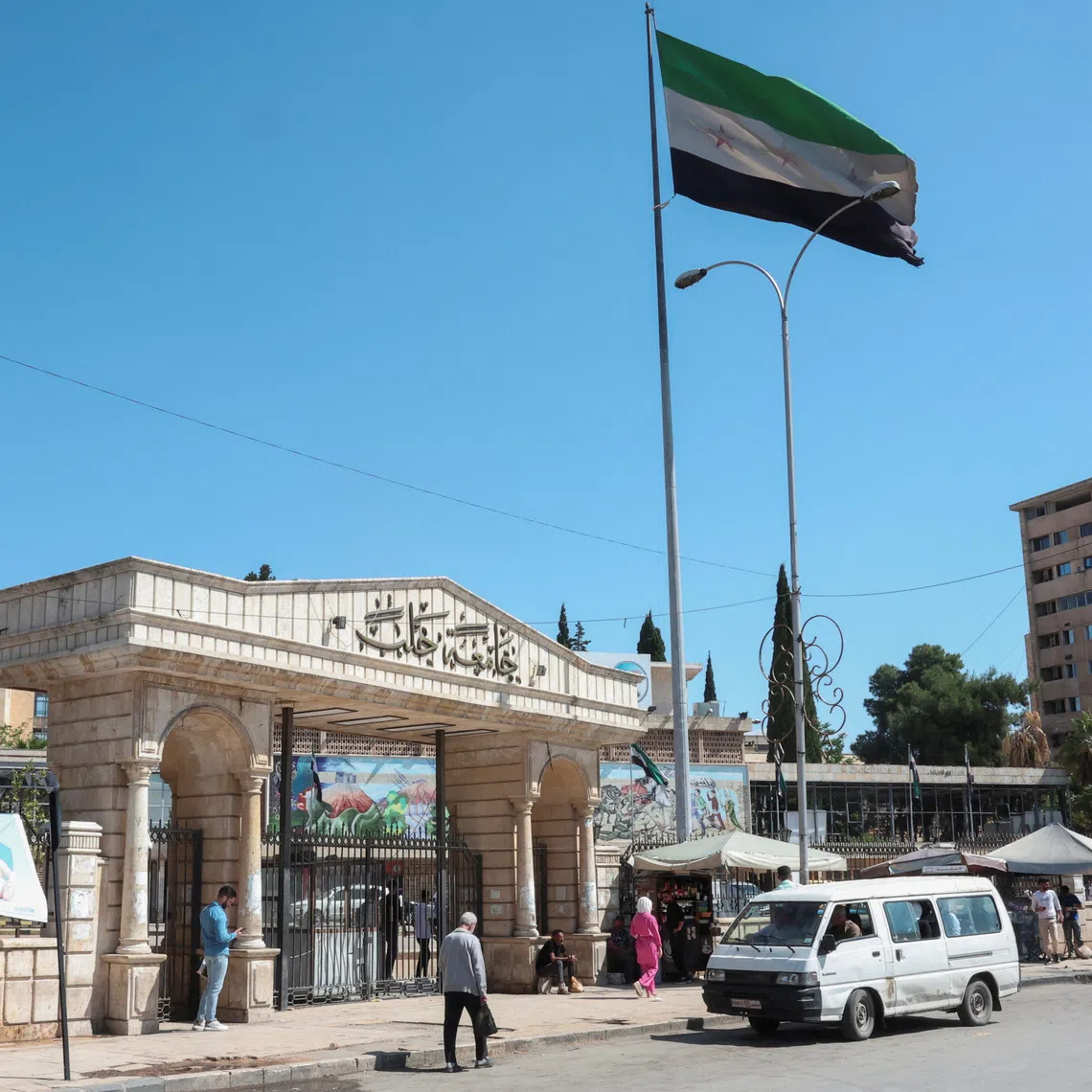 People walk near the Syrian flag, as U.S. President Donald Trump announced that he would order the lifting of sanctions on Syria, in Aleppo, Syria May 14, 2025. REUTERS/Mahmoud Hassano