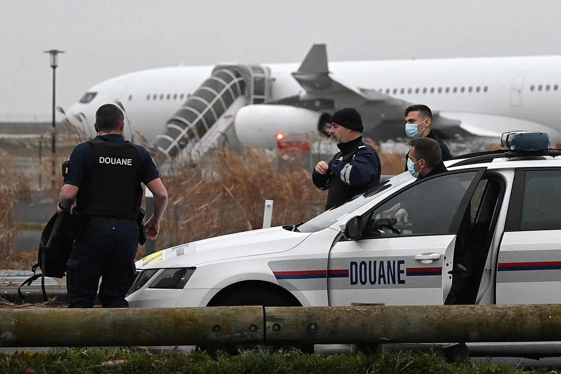 TOPSHOT - French customs officers stand next to a customs car with an Airbus A340 in the background which was grounded on the tarmac since December 21 over suspected "human trafficking", at the Vatry airport, north-eastern France on December 25, 2023. Two Indians passengers on the flight grounded since December 21, in the Marne region of France on suspicion of human trafficking will be presented to an examining magistrate on December 25, with possible indictment, while most of the other passengers are due to take off for India later on December 25. (Photo by FRANCOIS NASCIMBENI / AFP)