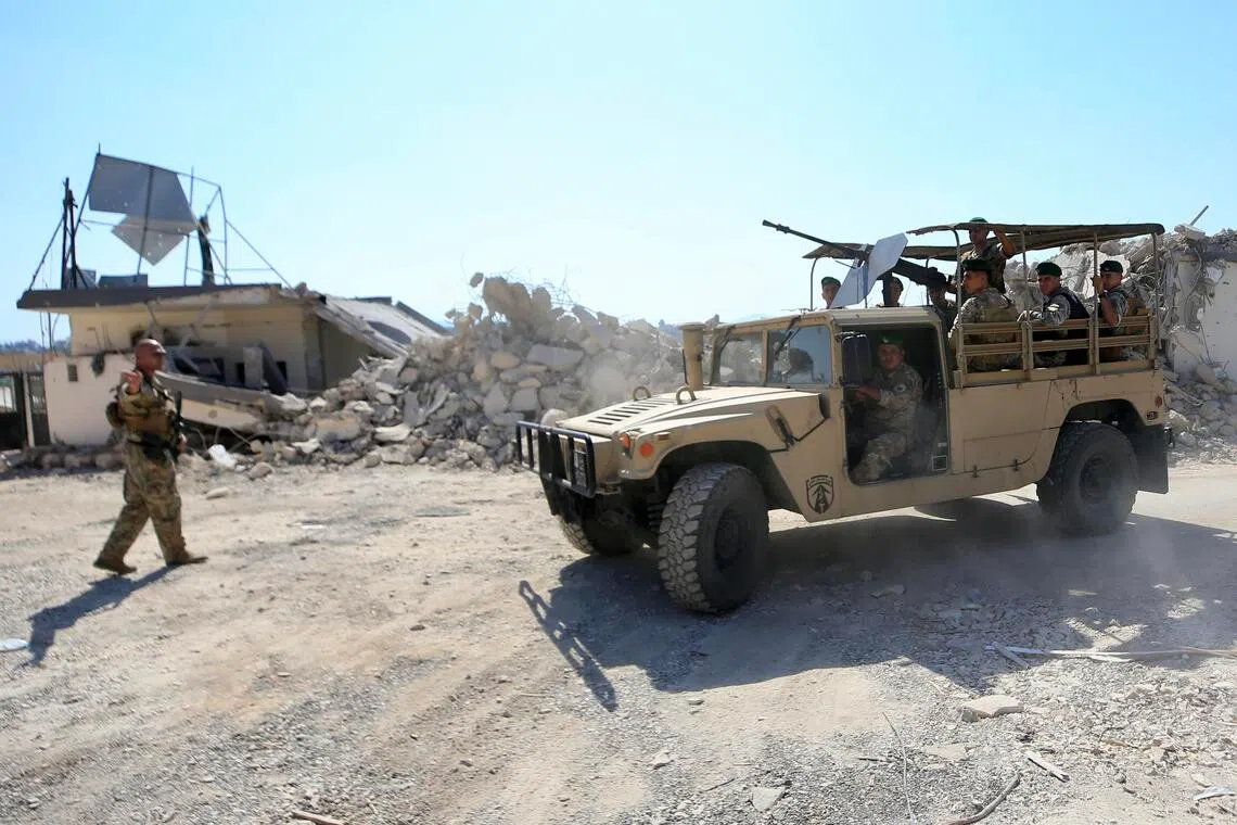 Military personnel stand near a tan tactical vehicle next to a damaged building.