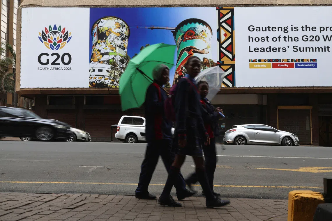 Schoolchildren holding umbrellas walk across the road from a G20 banner as South Africa prepares to host the G20 Summit on November 22-23, in Johannesburg, South Africa, November 18, 2025. REUTERS/Siphiwe Sibeko
