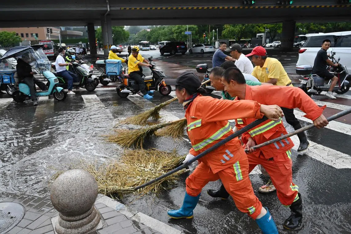 Workers sweeping water as scooter riders look on after heavy rain in Beijing on Saturday. 