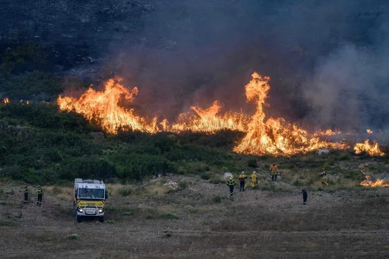 Wildfire burning in a field with emergency personnel and a vehicle nearby.