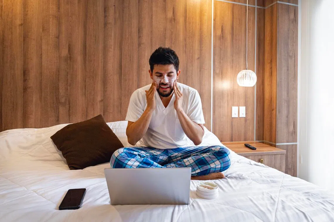 ANSKIN11 - Istockphoto - Young man sitting on bed in his bedroom, applying facial cream while watching a personal care tutorial on his laptop. Promotes skincare, wellness, beauty, self-care, and healthy lifestyle. Ideal for beauty routines, cosmetics, online learning, and modern home life concepts.