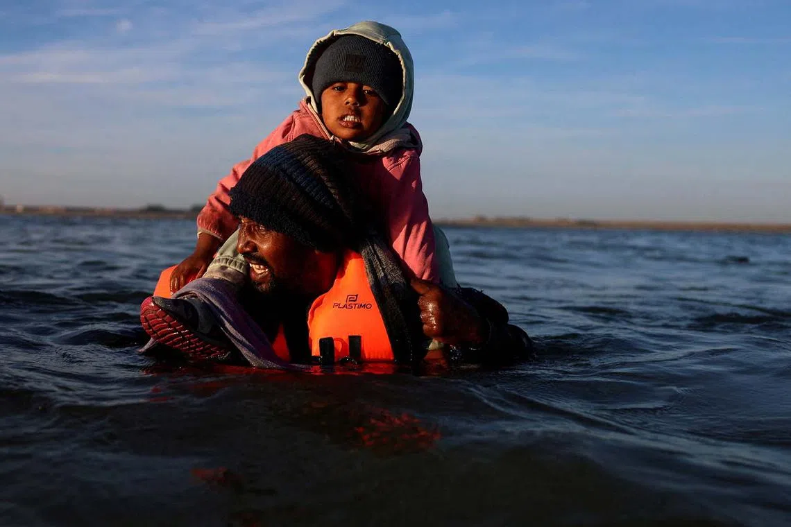 A migrant carrying a child wading through the sea in an attempt to board an inflatable dinghy that reaches Britain from the beach of Petit-Fort-Philippe in Gravelines, France, Aug 25, 2025. 