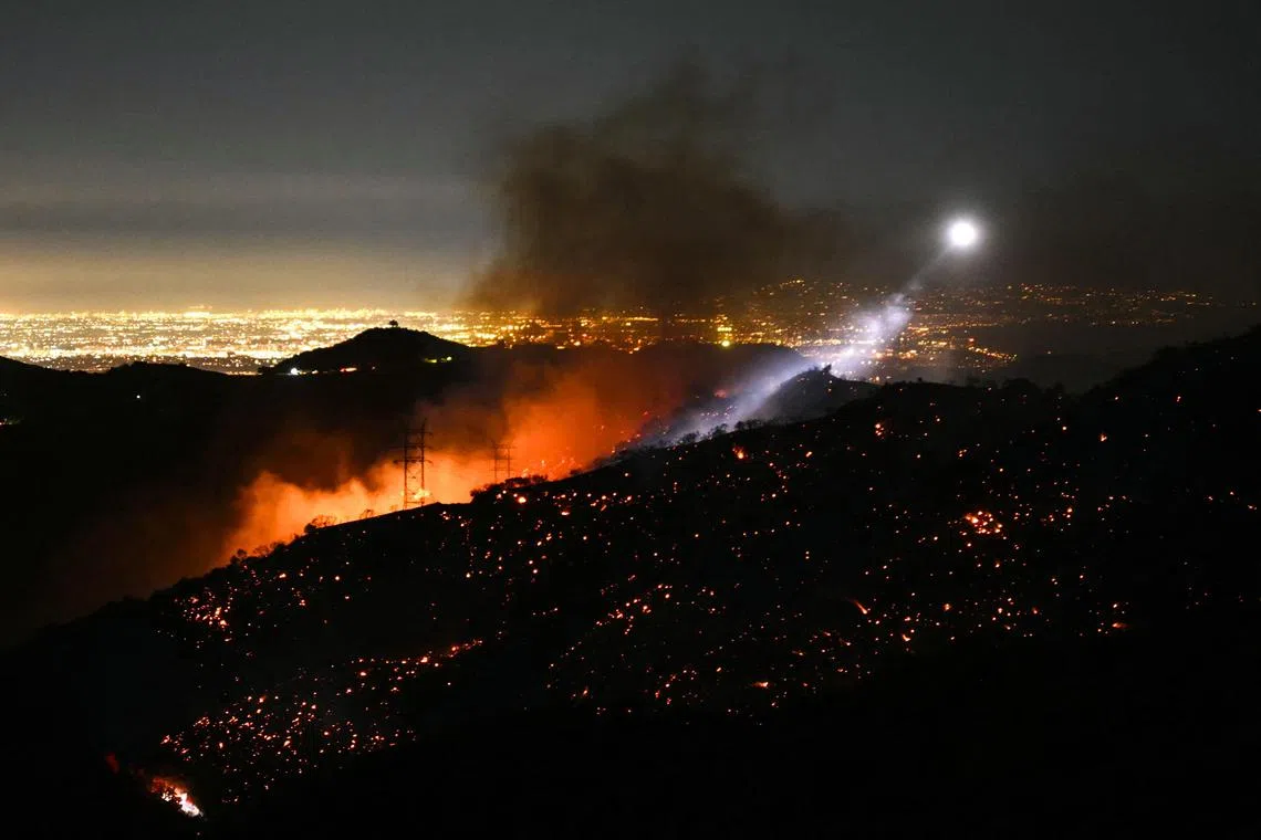A fire fighting helicopter illuminates a smouldering hillside as the Palisades fire grows near the Mandeville Canyon neighborhood and Encino, California, on Jan 11, 2025. 