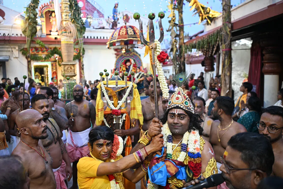 Volunteers portraying Sri Drowpathai (in yellow) and Sri Krishnan (wearing a crown) hold the battle flag and trident — symbols of the Kurukshetra battlefield — as they march to the Sri Drowpathai sanctum at Sri Mariamman Temple in the morning before preparations for the fire-walking ritual begins.