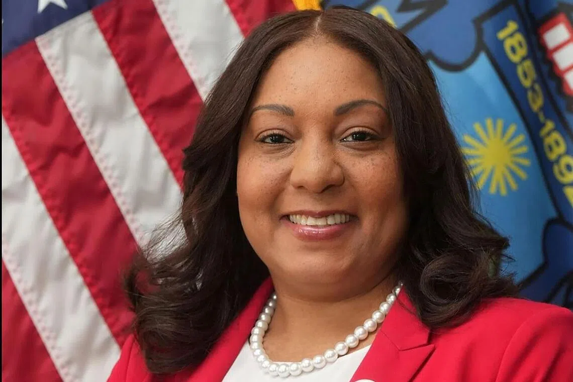 A smiling woman in a red blazer stands between the American flag and another flag.