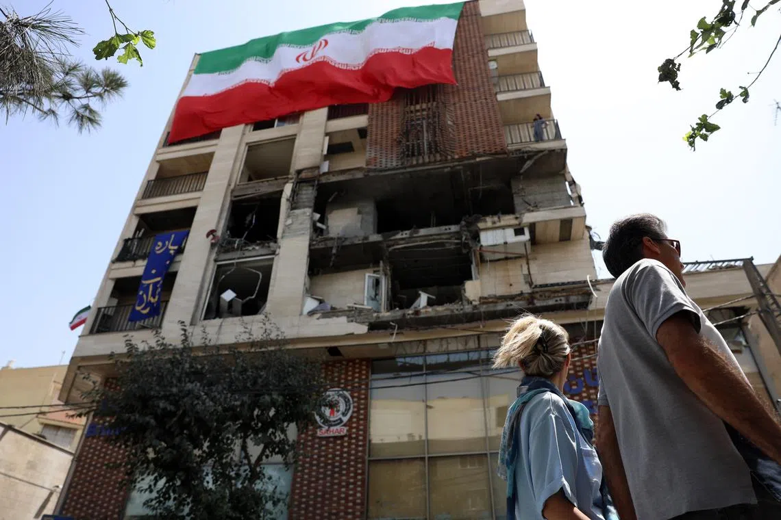 An Iranian couple walking past a national flag hanging from a damaged building in Tehran on June 25, following an Israeli airstrike during the 12-day war.