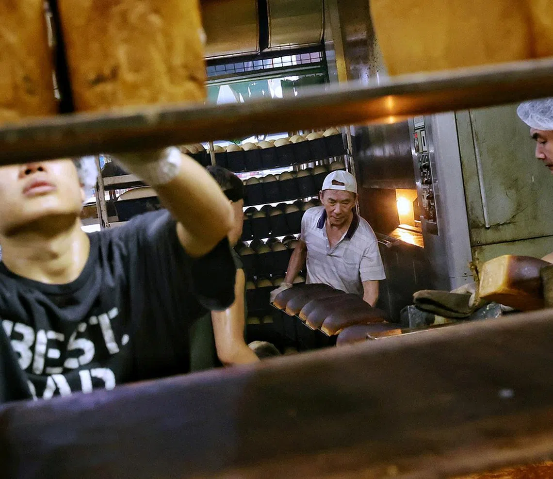 12. (SUGGESTED MAIN PIC 1) Senior baker Goh Wah Heng (centre), 72, taking out baking tins of traditional bread loaves from the revolving oven for his fellow bakers to load onto a rack to cool in the kitchen of Sweetlands Confectionery and Bakery in Kim Keat Lane on May 15, 2025. The bakery can produce more than 1,000 loaves of traditional bread in a day.