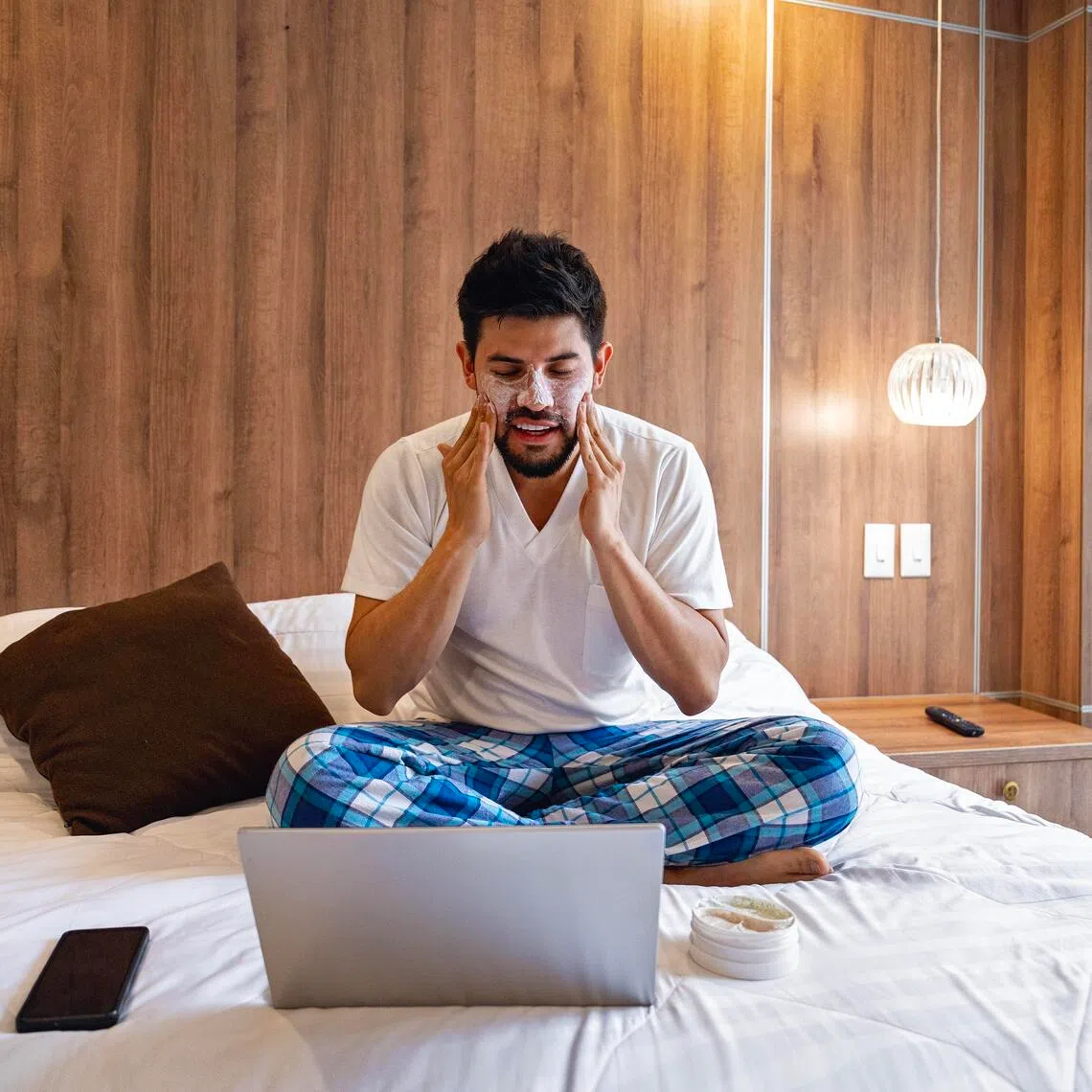 ANSKIN11 - Istockphoto - Young man sitting on bed in his bedroom, applying facial cream while watching a personal care tutorial on his laptop. Promotes skincare, wellness, beauty, self-care, and healthy lifestyle. Ideal for beauty routines, cosmetics, online learning, and modern home life concepts.