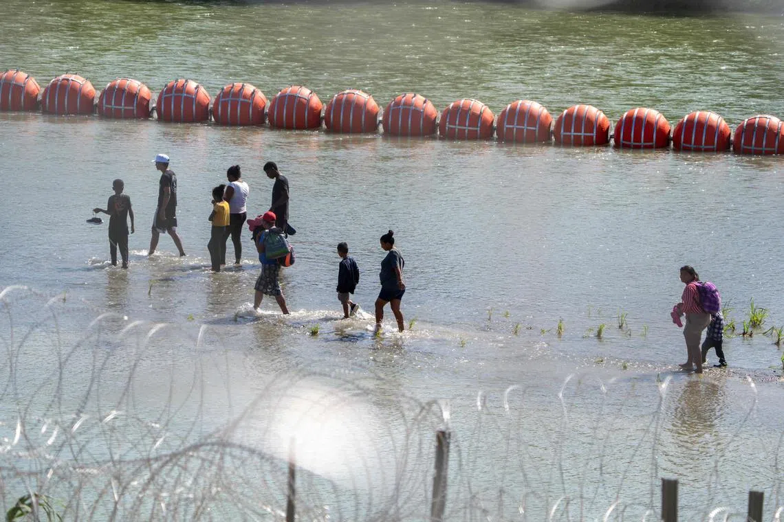 Migrants walk past a string of buoys placed on the water along the Rio Grande border with Mexico in Eagle Pass, Texas, on July 15.