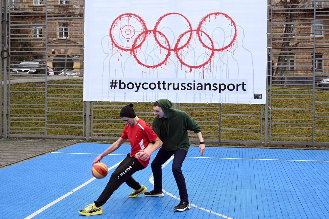 A banner featuring bloodied Olympic rings and the hashtag #boycottssiansport hangs on the fence of an outdoor sports ground while boys play basketball in the western Ukrainian city of Lviv.