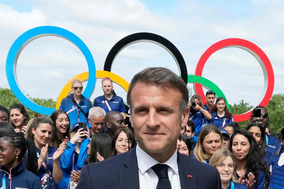 French President Emmanuel Macron during a visit to the Athletes' Village with the Olympic rings displayed behind him and French athletes in the background.