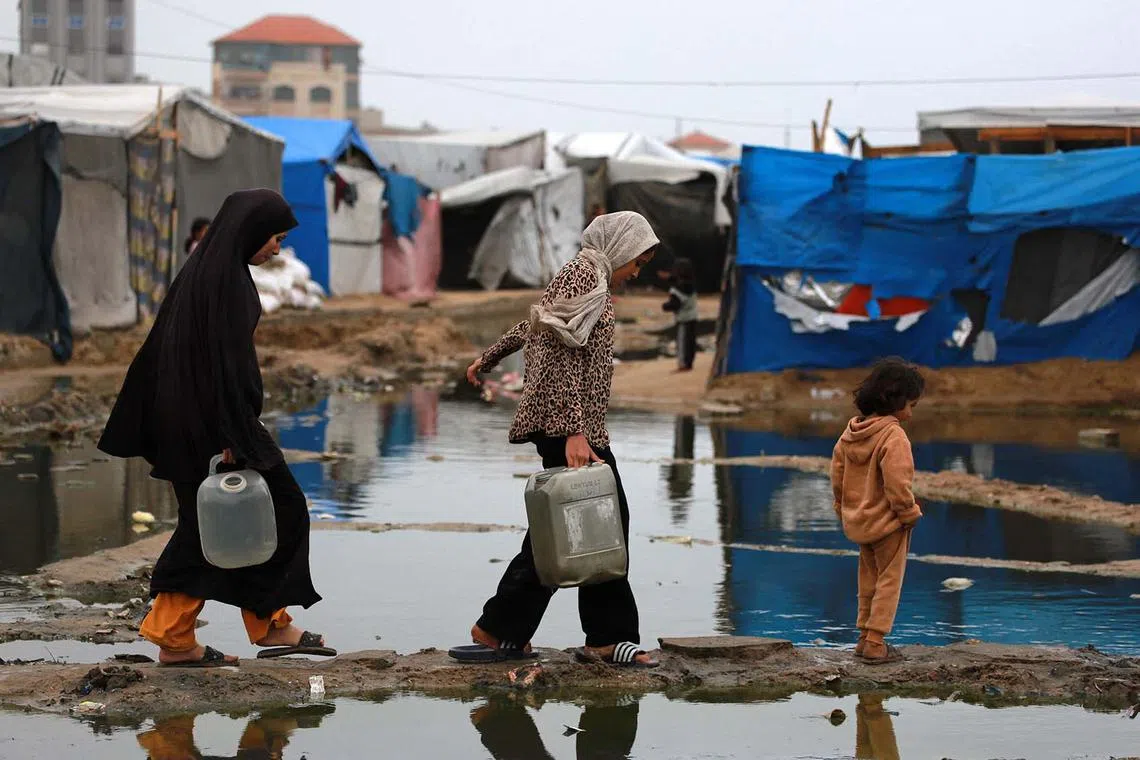 Displaced Palestinian women using a dry path to carry water to their tent, following heavy rainfall north of Deir el-Balah in the central Gaza Strip on Nov 24, 2024, amid the ongoing war between Israel and Hamas militants. 