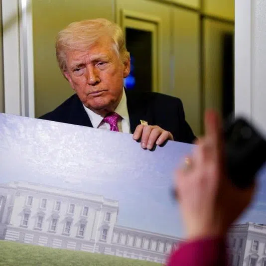 US President Donald Trump speaking to the media while holding up renderings of the planned White House ballroom, aboard Air Force One on March 29, 2026.