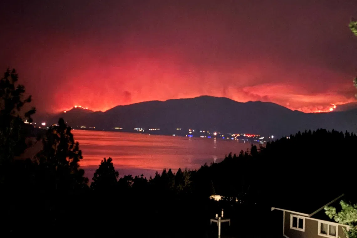 Night view of a body of water reflecting a large fire burning on distant hills.