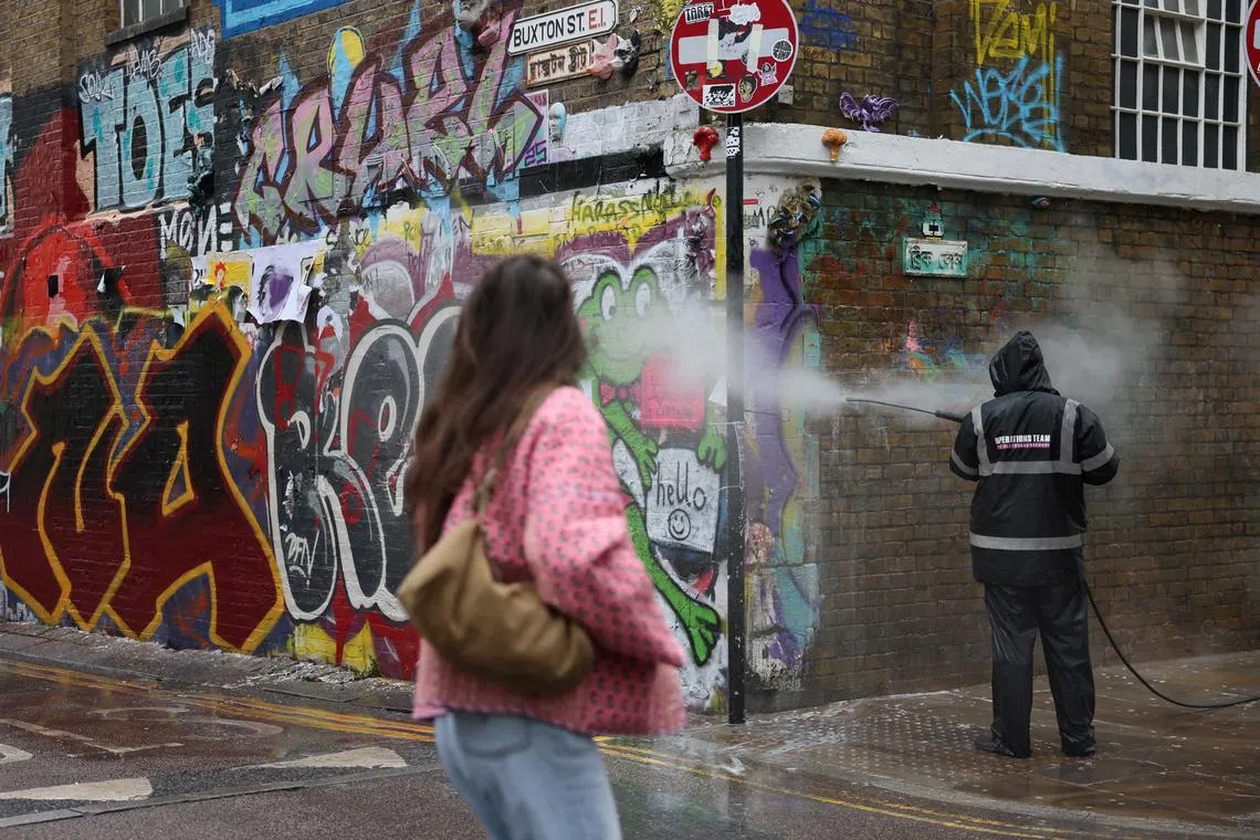 FILE PHOTO: A woman walks past a worker cleaning graffiti from a wall in Brick Lane, east London, Britain, March 25, 2025. REUTERS/Isabel Infantes/File Photo