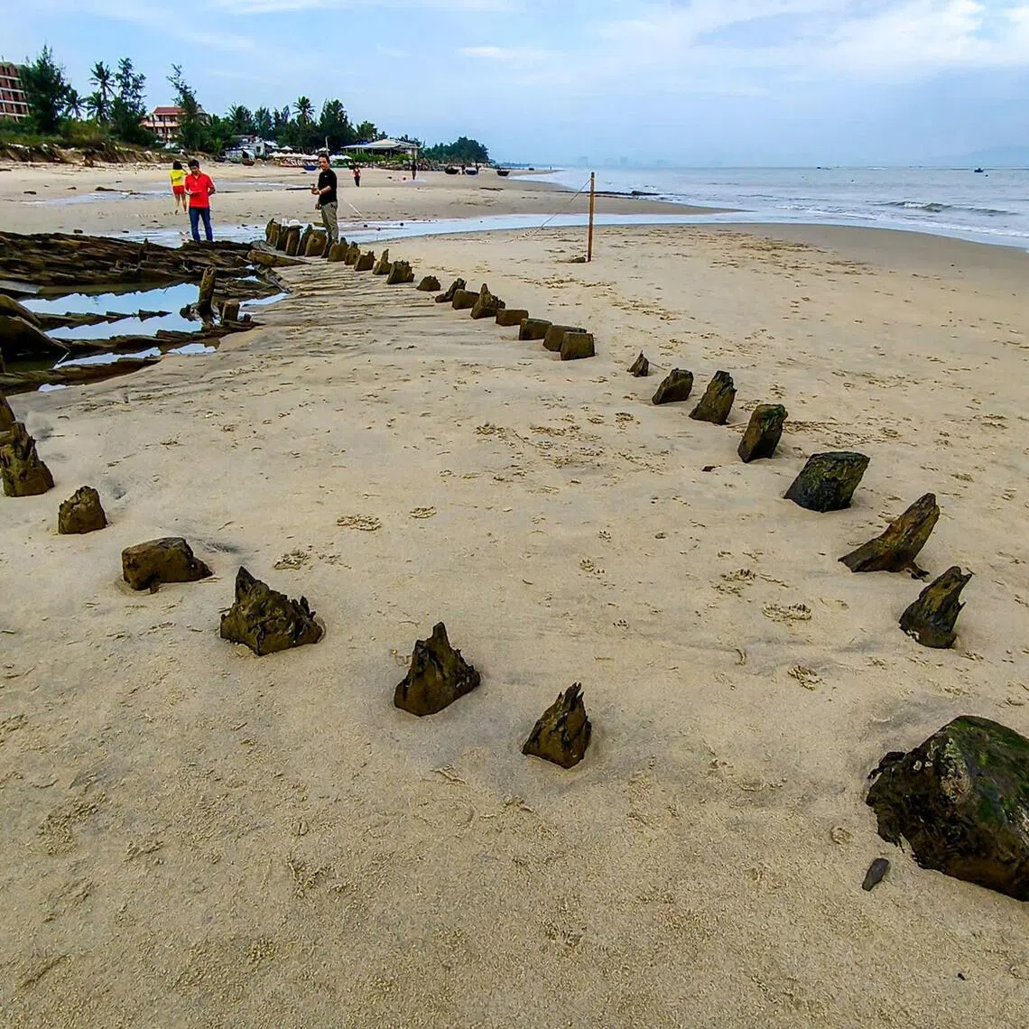 People stand next to a centuries-old shipwreck uncovered in the aftermath of Typhoon Kalmaegi on a beach off the Hoi An coast in central Vietnam, on November 10, 2025. Initially discovered in 2023, the at least 17.4-metre (57-foot) vessel -- whose heavy wood-ribbed hull survived hundreds of years of rough seas almost perfectly intact -- was resubmerged before authorities could reclaim it. (Photo by Tam Xuan / AFP)